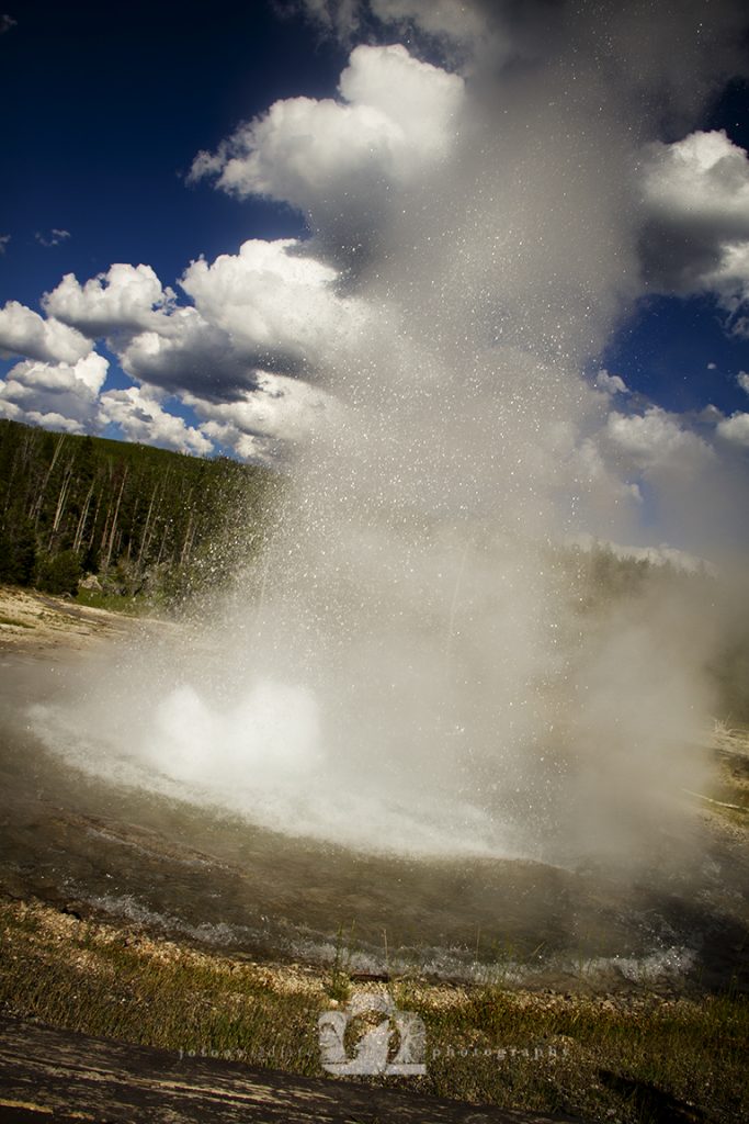 An erupting geyser in Yellowstone with a big spray of water drops captured against a blue sky with white clouds